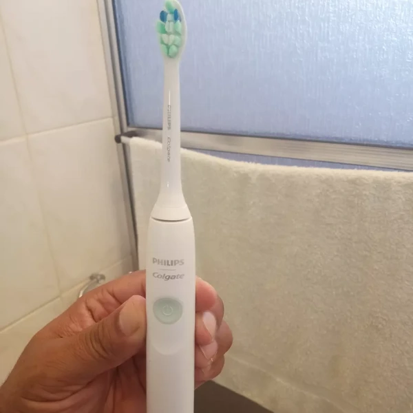 Escova Philips Colgate SonicPro 10 sendo segurada por uma mão no banheiro com toalha ao fundo
